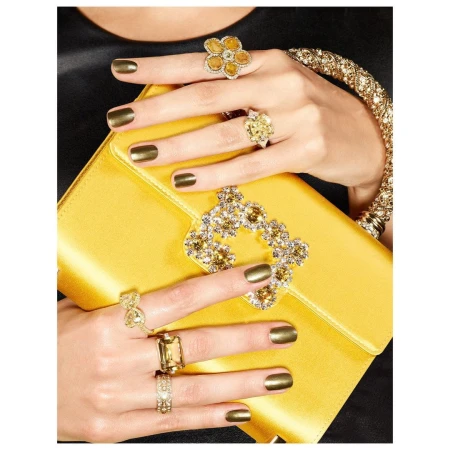 Close-up of hands displaying gold gemstone rings and metallic olive nail polish while holding a yellow satin handbag.