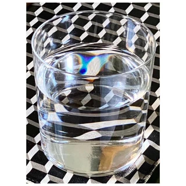 Clear glass filled with water sitting on a black and white geometric patterned surface, creating refracted light patterns and reflections.