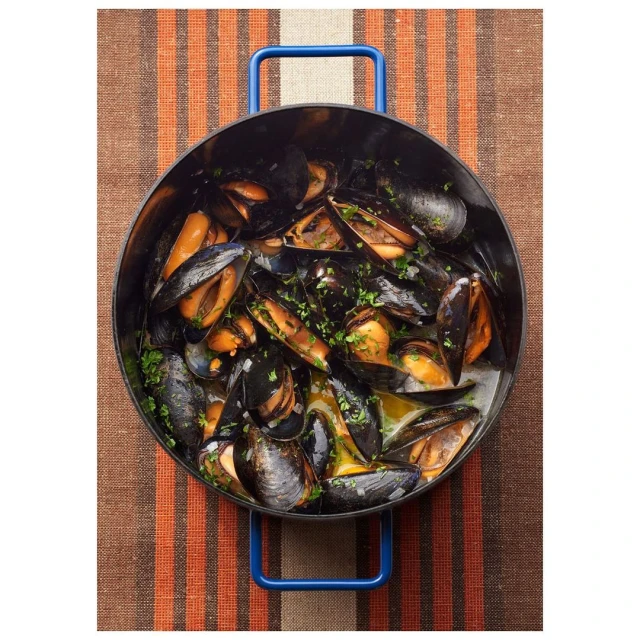 Overhead shot of cooked mussels in a black pot with blue handles, garnished with parsley, sitting on a brown and orange striped placemat.