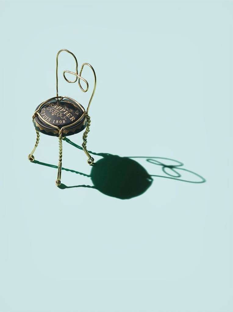 A whimsical miniature chair crafted from a champagne bottle cap and delicate gold wire stands against a soft, light-blue backdrop.