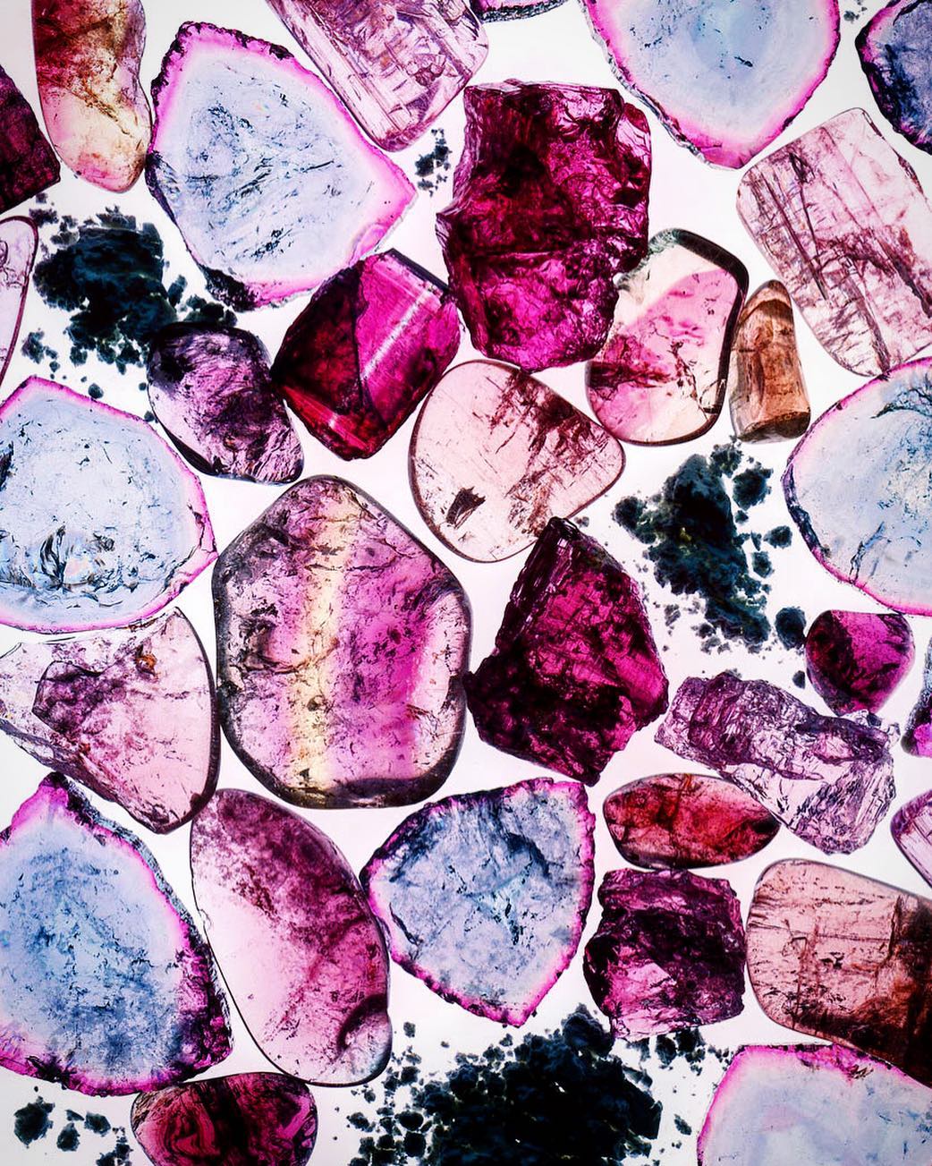 Macro photograph of translucent mineral grains with pink, blue, and purple hues. Grains are scattered against a bright white background.