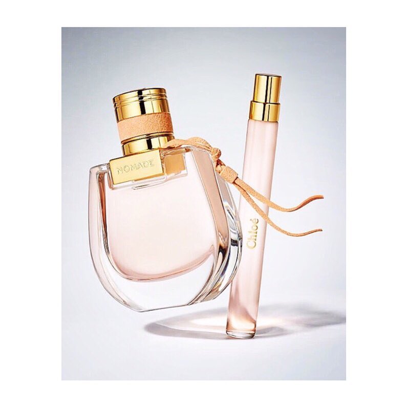 Two perfume bottles, one round with a beige strap and a smaller rectangular bottle, stand on a light surface with soft, diffused lighting.