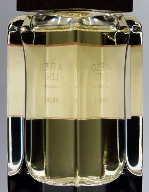 Close-up of a clear glass perfume bottle with a dark brown cap, filled with light yellow liquid, and featuring Spanish text on the label.