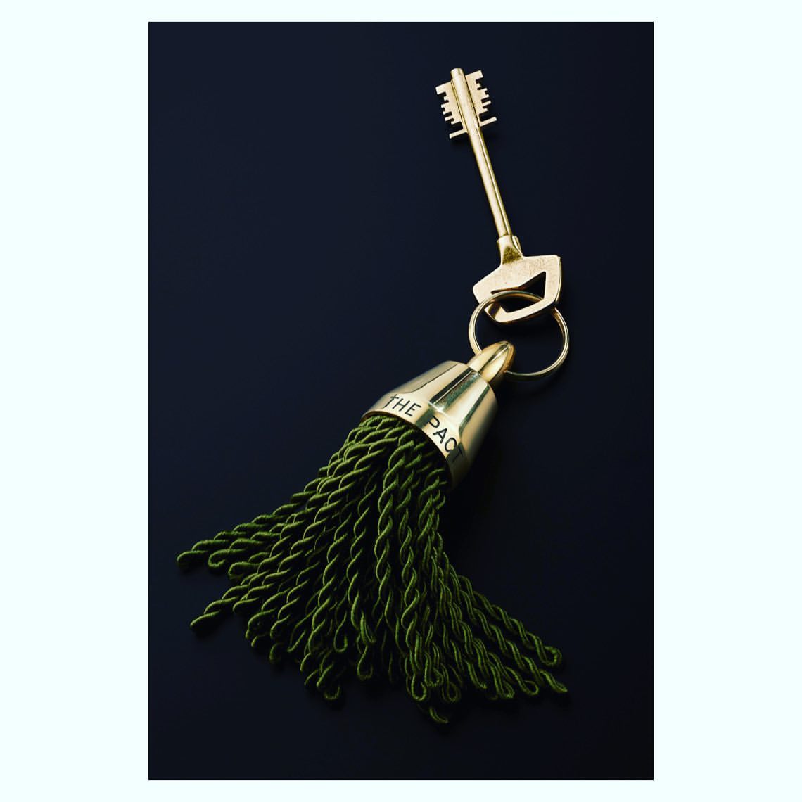 Stylish gold key with green tassel on a dark background. Key ring and cap are metallic gold, with dark green tassel threads.