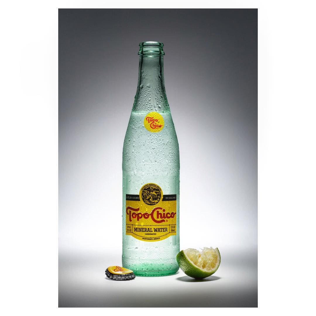 A bottle of Topo Chico mineral water stands with a lime wedge and bottle cap against a grey gradient backdrop in a studio shot.