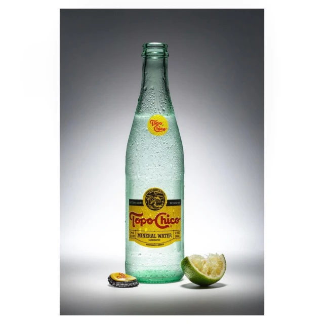 A bottle of Topo Chico mineral water stands with a lime wedge and bottle cap against a grey gradient backdrop in a studio shot.