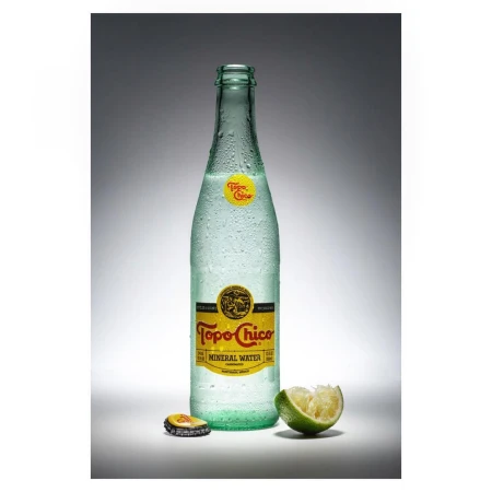 A bottle of Topo Chico mineral water stands with a lime wedge and bottle cap against a grey gradient backdrop in a studio shot.