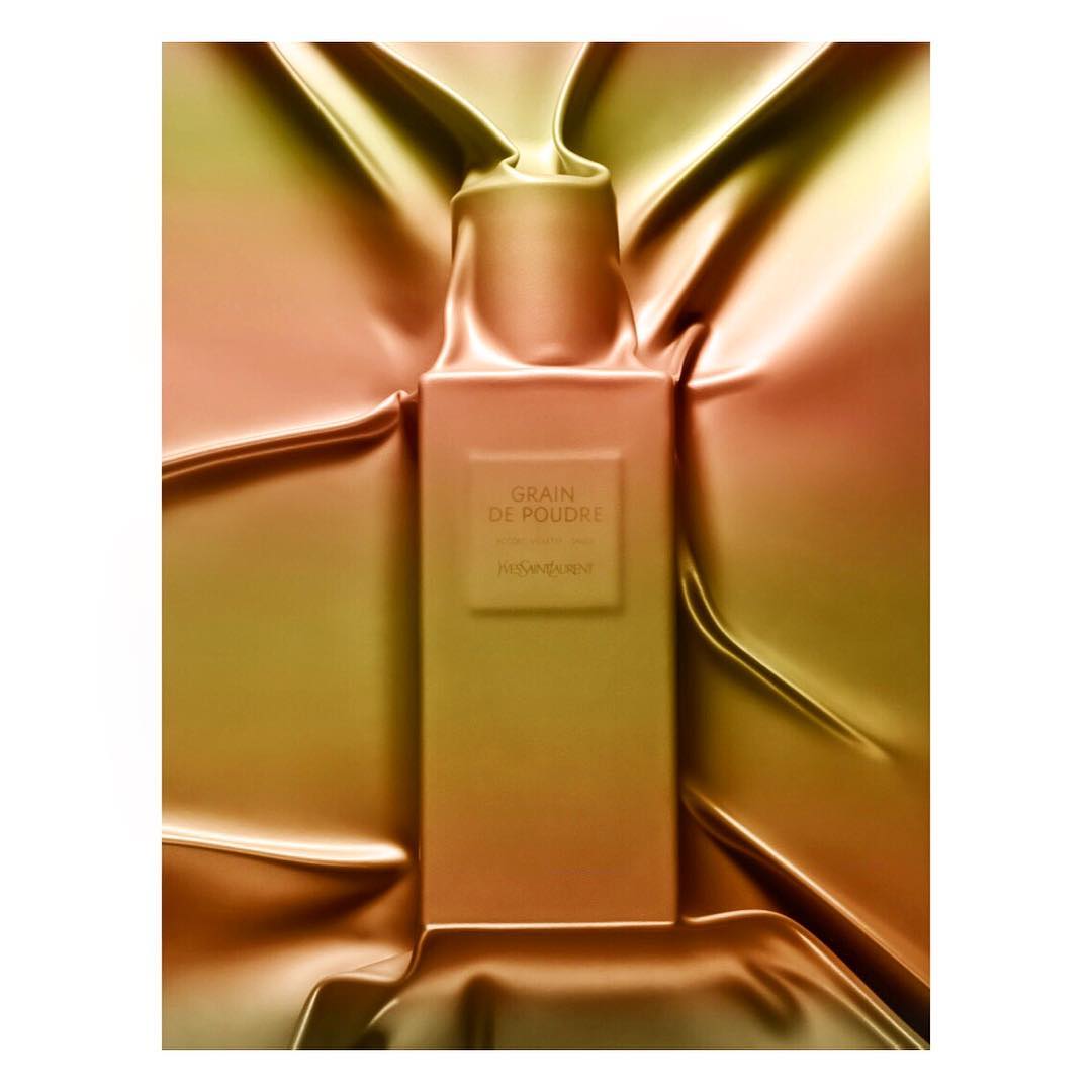Close-up of a rectangular YSL Grain de Poudre perfume bottle surrounded by draped fabric in shades of gold, brown, and peach.