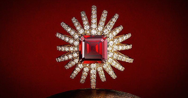 Close-up of a gold ring featuring a large, square-cut red ruby surrounded by numerous small diamonds, set against a red backdrop.