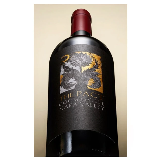 A black wine bottle with an ornate gold and white label featuring “The Pact” over a tan background is presented with a low-angle perspective.