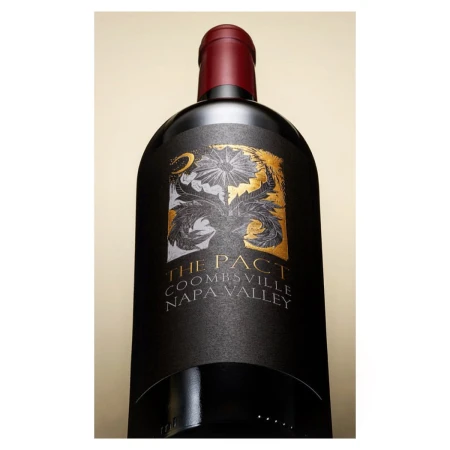 A black wine bottle with an ornate gold and white label featuring “The Pact” over a tan background is presented with a low-angle perspective.