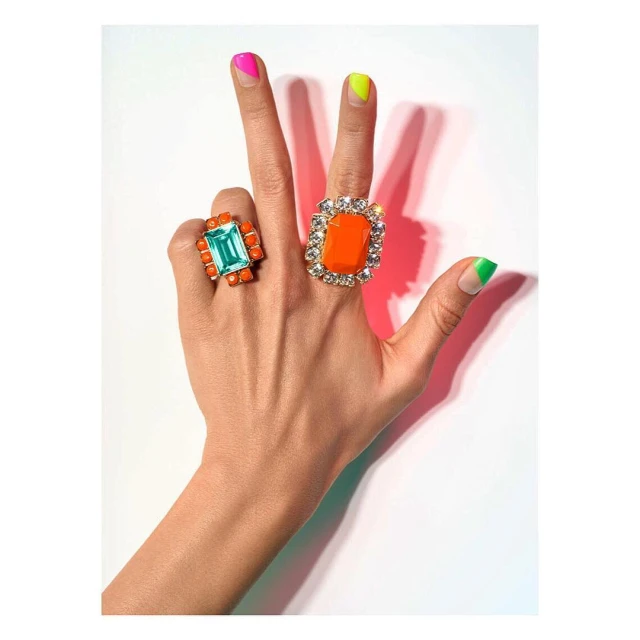 Woman's hand displays bright manicured nails and two statement rings with colorful gemstones against a white background in a beauty shot.