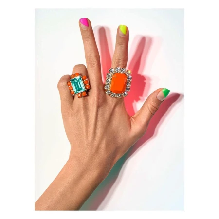 Woman's hand displays bright manicured nails and two statement rings with colorful gemstones against a white background in a beauty shot.