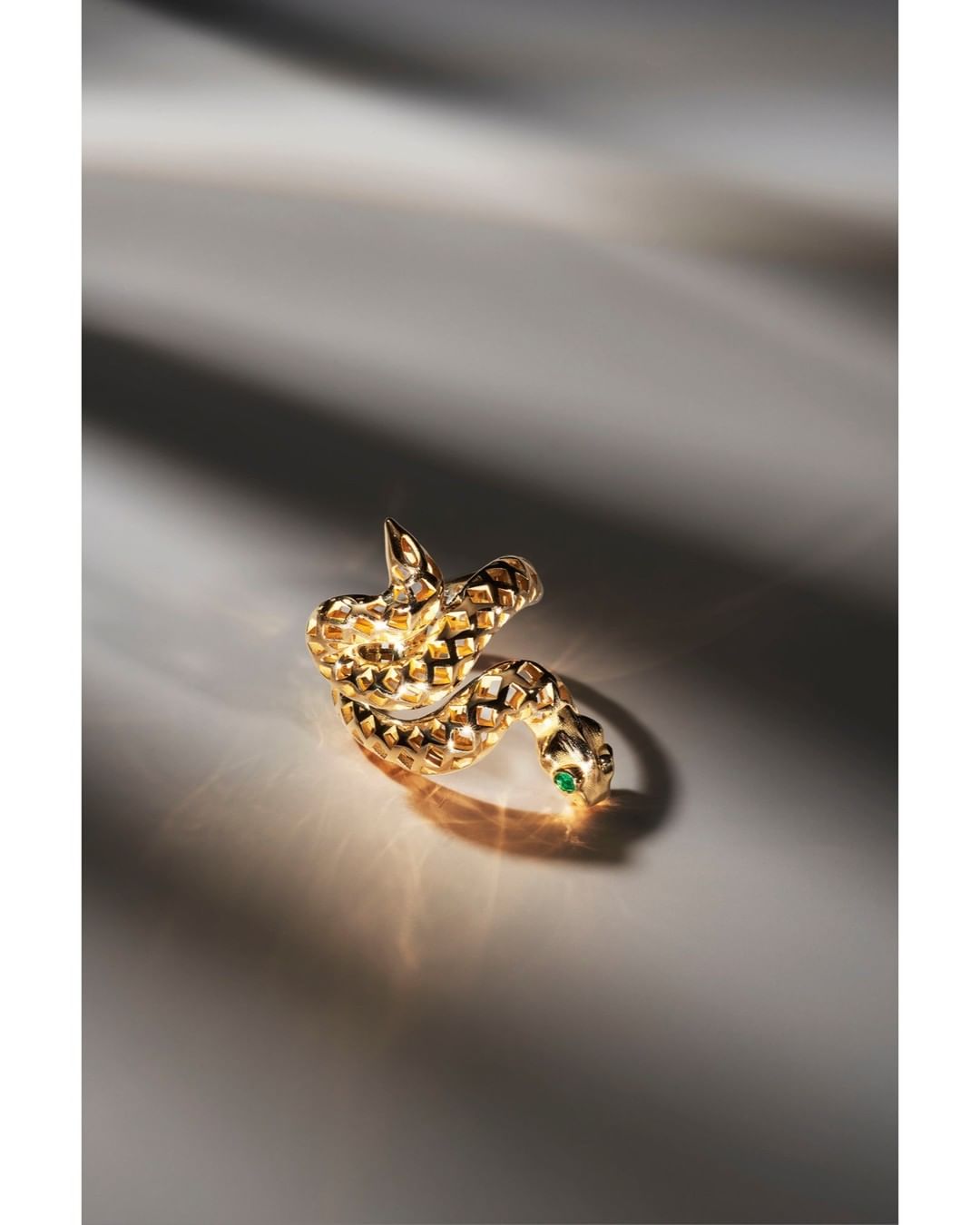 Close-up shot of a gold snake ring with a green gemstone detail coiled on a neutral surface with soft, directional lighting.