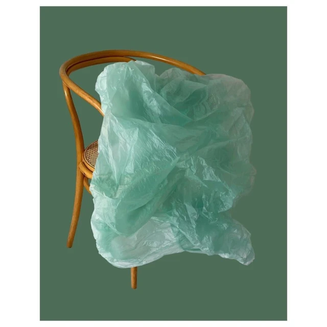 A tan bentwood chair is draped with a translucent, light-green plastic sheet, set against a solid dark-green backdrop in a studio setting.