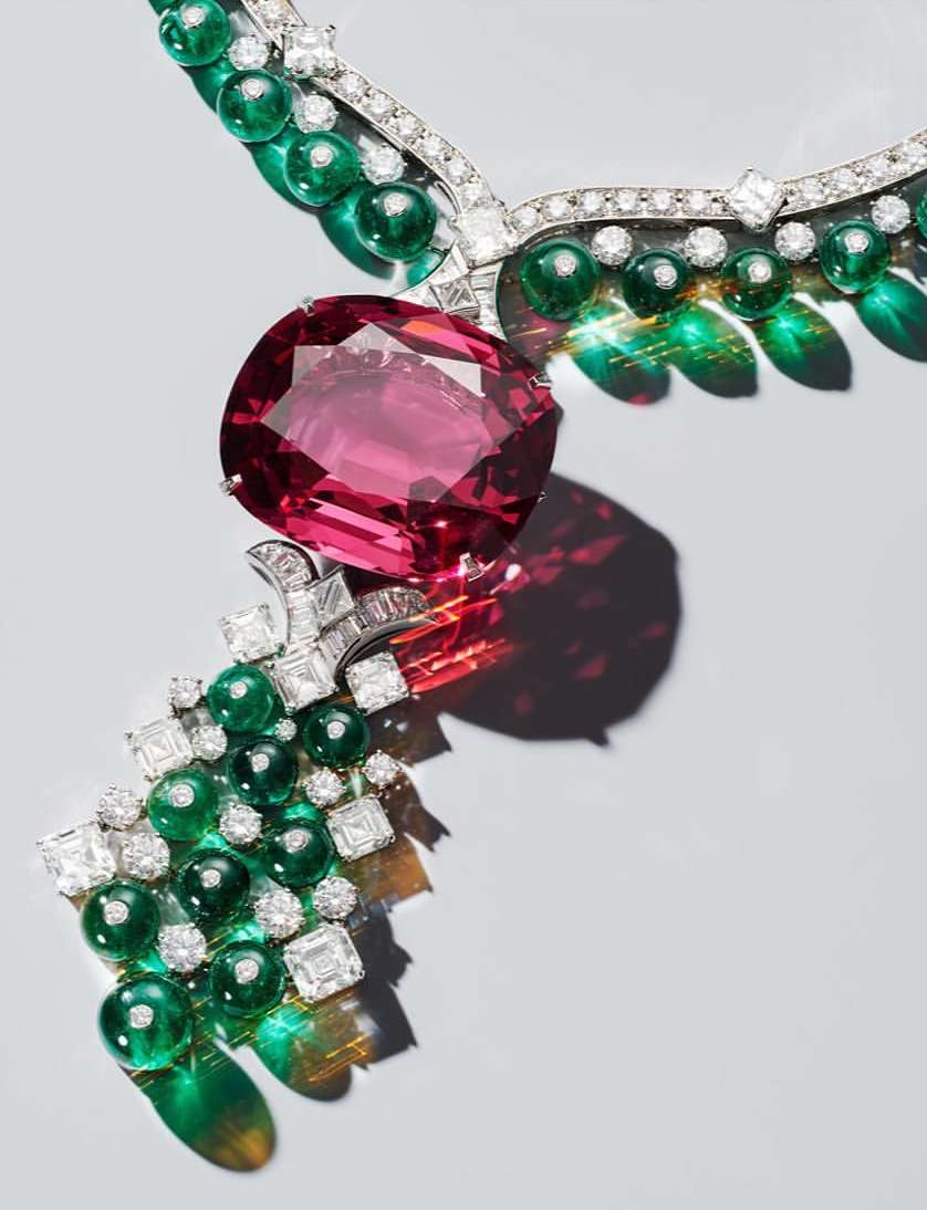 A close-up of a luxurious necklace featuring a large pink gemstone, emerald green stones, and diamonds against a soft grey backdrop.