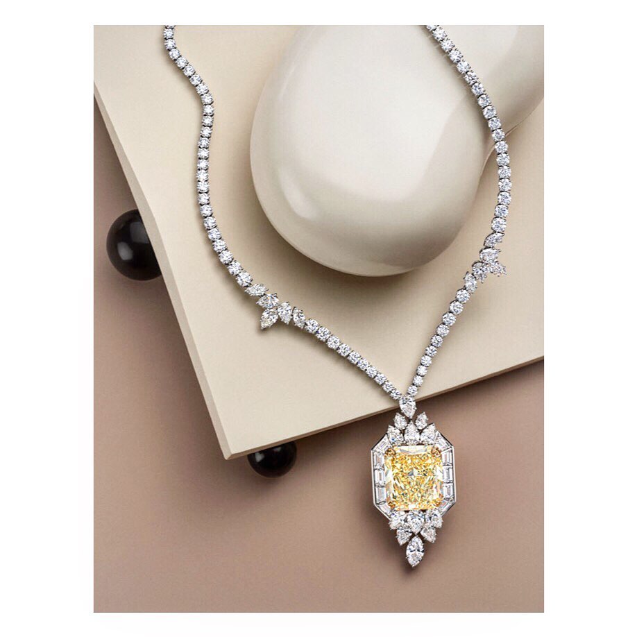 Close-up of an elegant diamond necklace featuring a large yellow diamond pendant, set against a modern neutral background with decorative orbs.