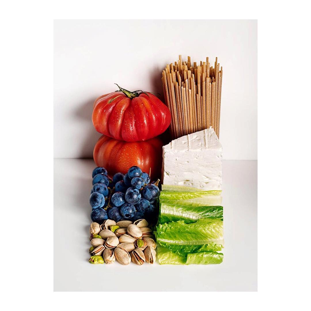 Overhead shot of organized food ingredients: tomatoes, pasta, feta cheese, grapes, lettuce, pistachios on white background, food photography.