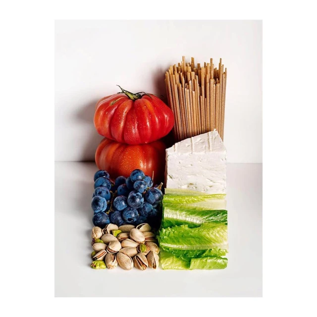 Overhead shot of organized food ingredients: tomatoes, pasta, feta cheese, grapes, lettuce, pistachios on white background, food photography.