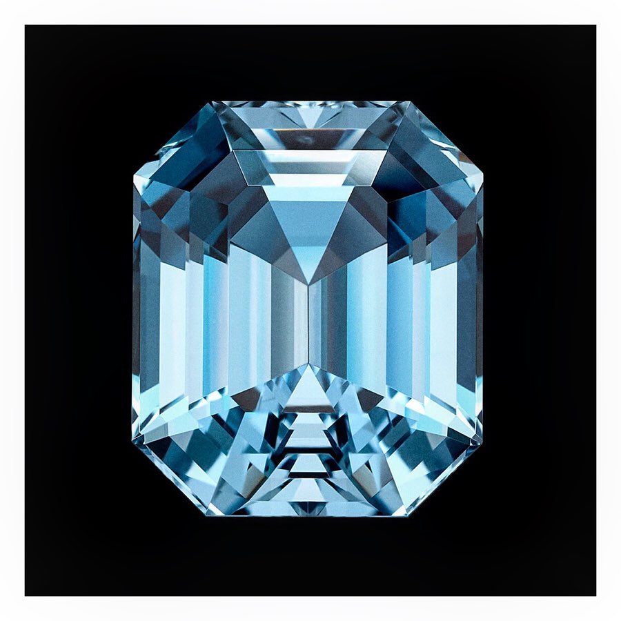 Close-up studio shot of a faceted, rectangular, light-blue gemstone with geometric details and reflections on a dark-black background.