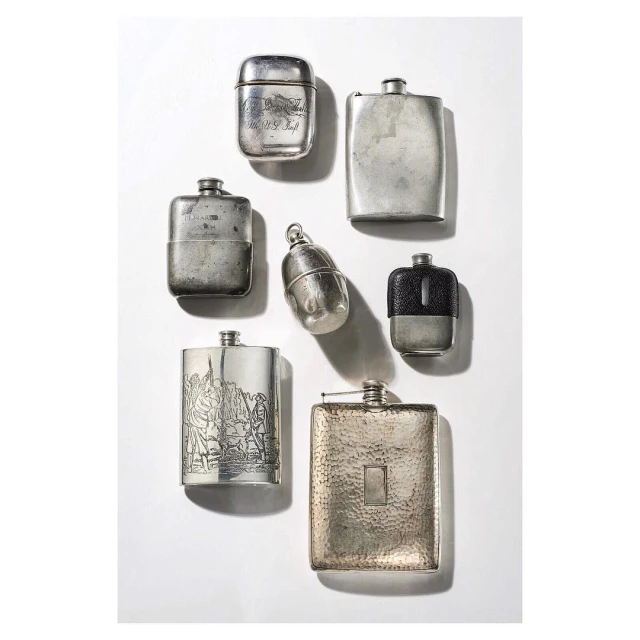 Still life shot of a collection of silver hip flasks of varying shapes and sizes arranged on a white background with shadows.
