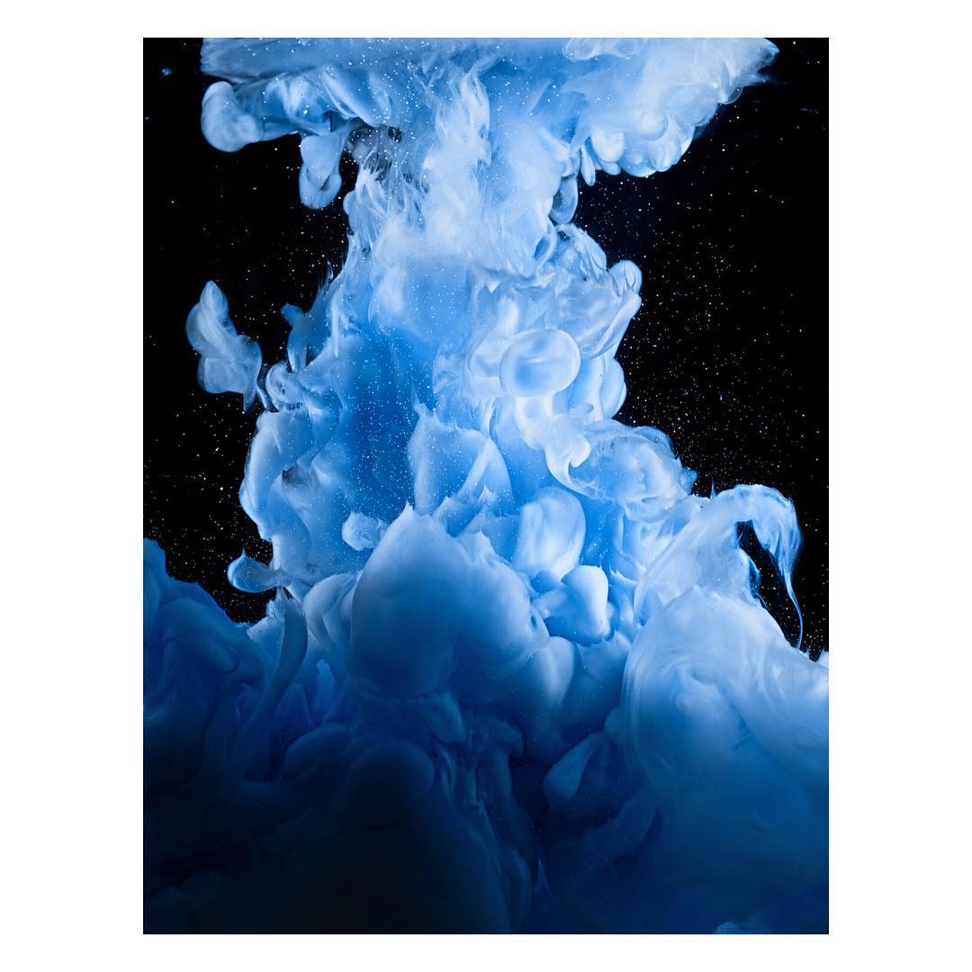Abstract image of blue and white ink swirling in water against a black background creating a cloud-like and ethereal effect.