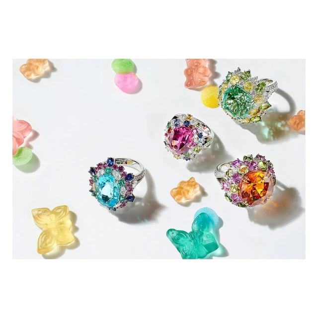 Jewellery still life featuring four gemstone rings with diamonds and colorful candy bears on a white surface in bright studio lighting.