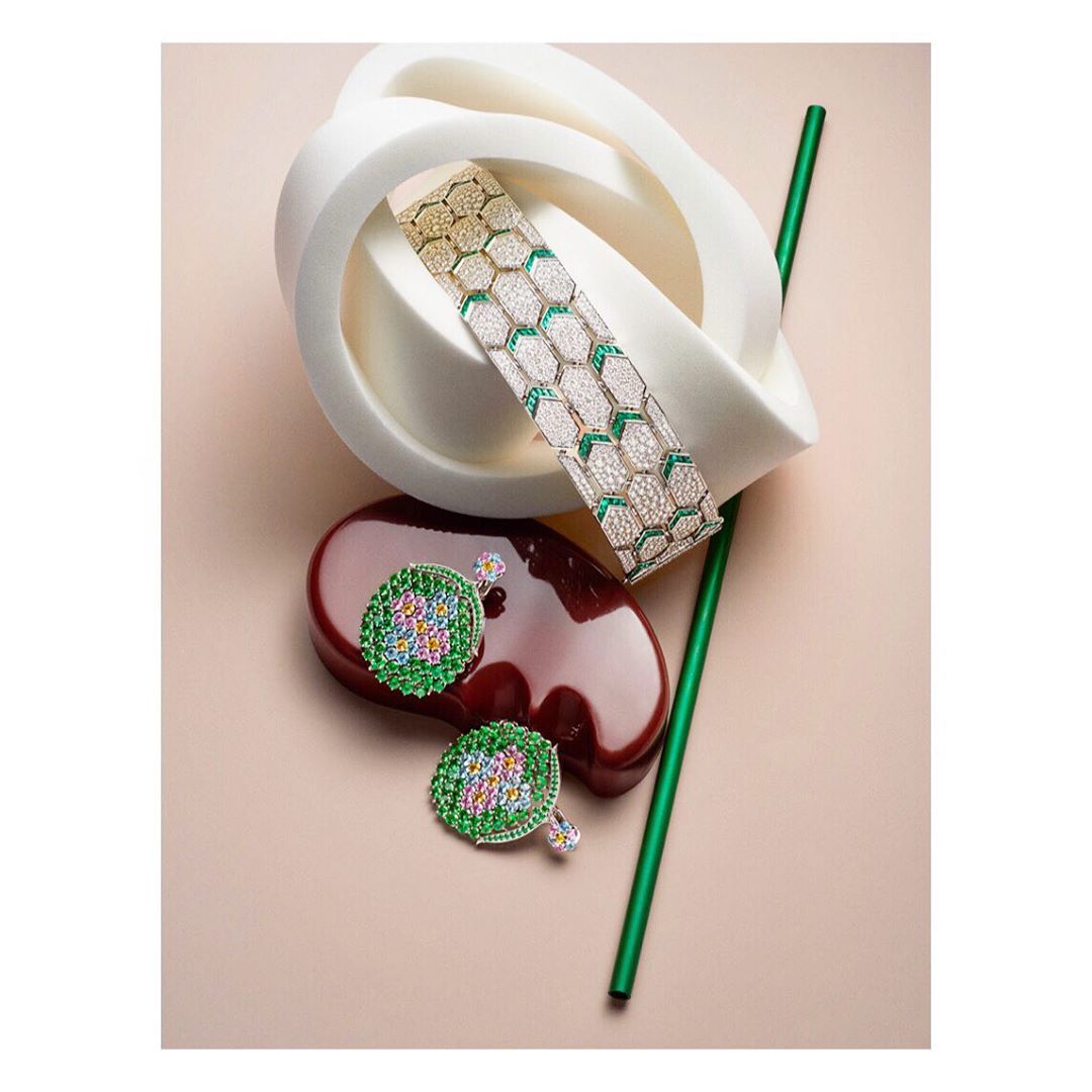 Still life shot of a gold bracelet with white and green gems, green-and-gold earrings, a green straw, and geometric shapes on a peach background.