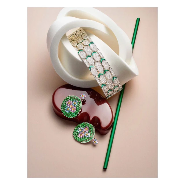 Still life shot of a gold bracelet with white and green gems, green-and-gold earrings, a green straw, and geometric shapes on a peach background.