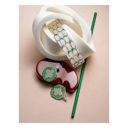 Still life shot of a gold bracelet with white and green gems, green-and-gold earrings, a green straw, and geometric shapes on a peach background.