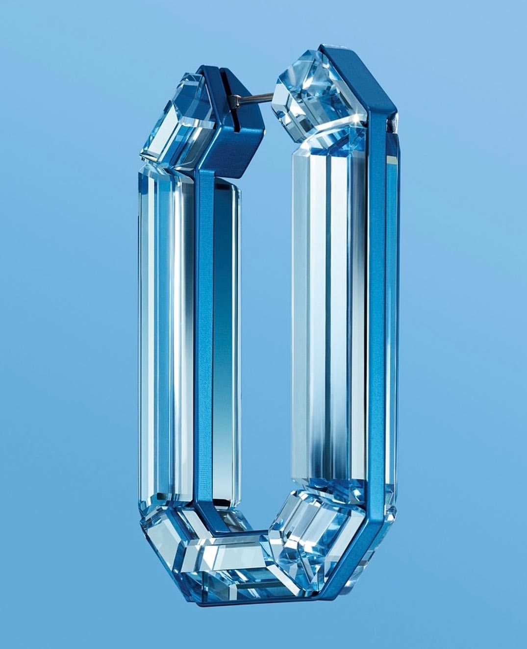 Close-up of modern blue and clear crystal hoop earring with multifaceted surface, studio shot against soft blue backdrop.