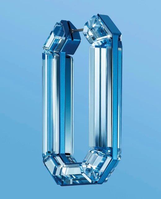 Close-up of modern blue and clear crystal hoop earring with multifaceted surface, studio shot against soft blue backdrop.
