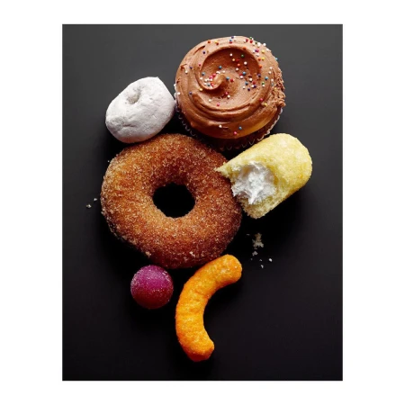 Overhead shot of an assortment of sweets and sugary snacks, including a frosted cupcake, sugar donut, Twinkie, and cheese puff, on a black surface.