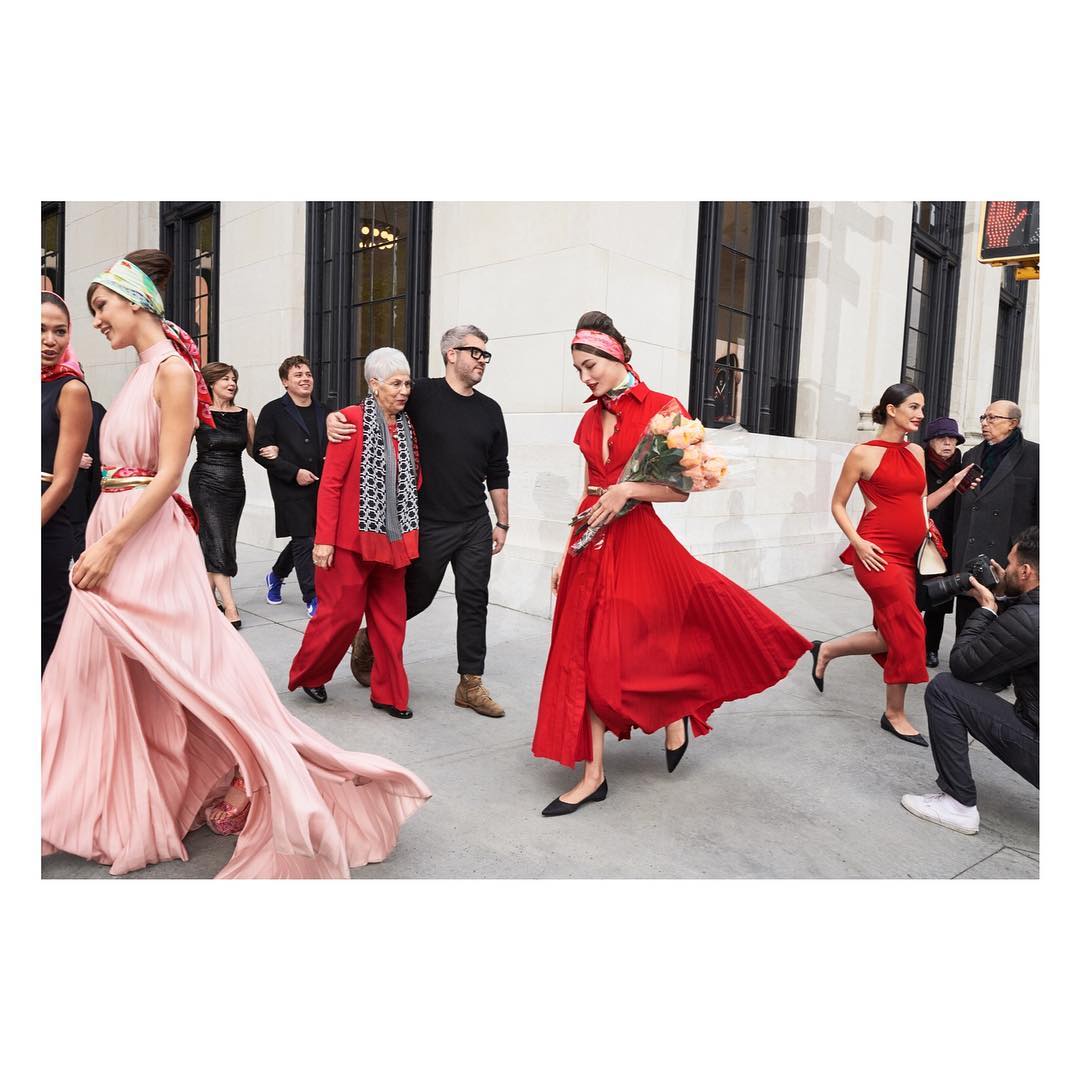 Several fashion models walk together on a city street, wearing colourful headscarves and red and pink pleated dresses and suits.