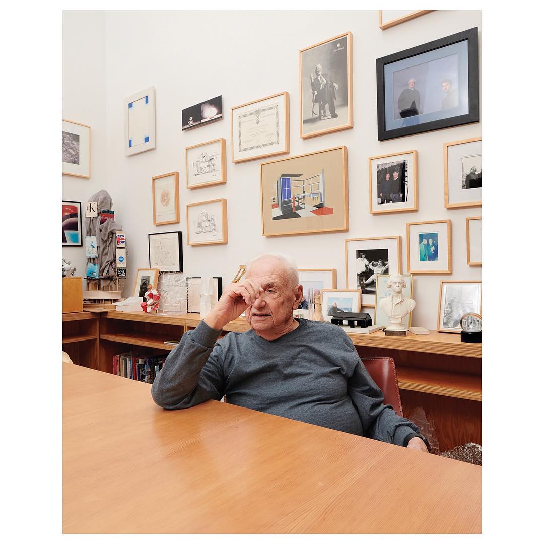 An older adult man is seated at a wooden table in an office setting with framed pictures hanging on the wall behind him.