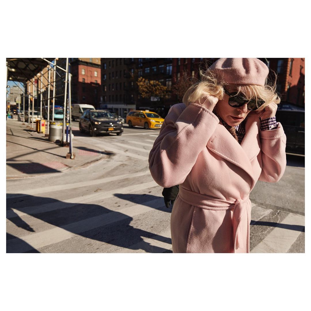 An adult woman with blonde hair, wearing a pink coat and beret, walks on a crosswalk in a city street with cars and buildings.