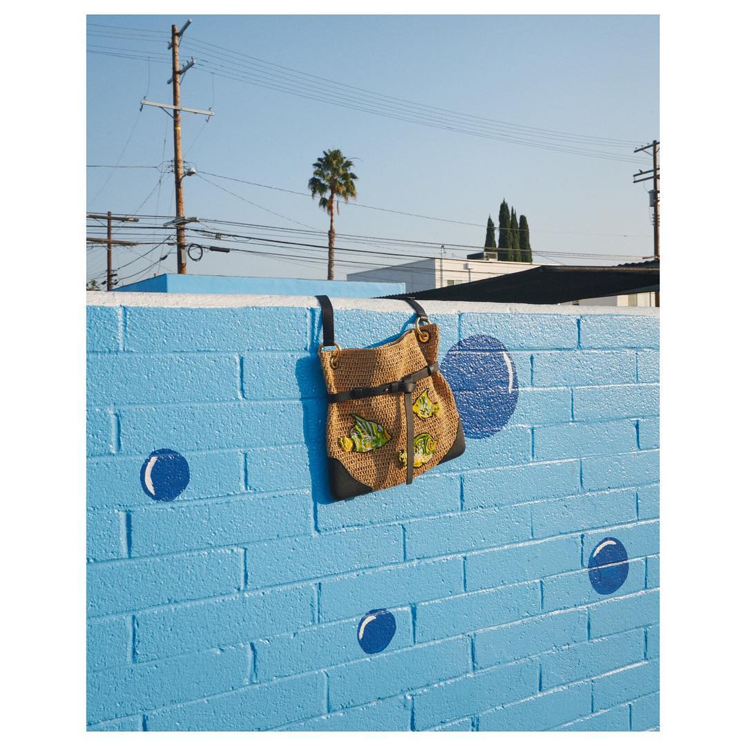 A jute purse with fish applique hangs from a bright blue brick wall with bubble painting, under a sunny sky with palm trees.