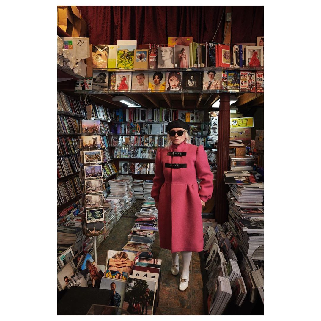 Blonde woman in a pink coat and black beret stands in a narrow shop aisle lined with records and magazines on shelves and the floor.