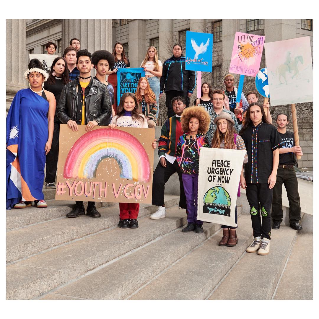 Group of youth activists stand on building steps holding signs with messages of protest and environmental awareness for a demonstration.