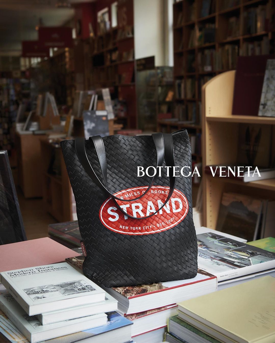 A black Bottega Veneta tote bag with a Strand Bookstore logo on a stack of books in a bookstore setting.