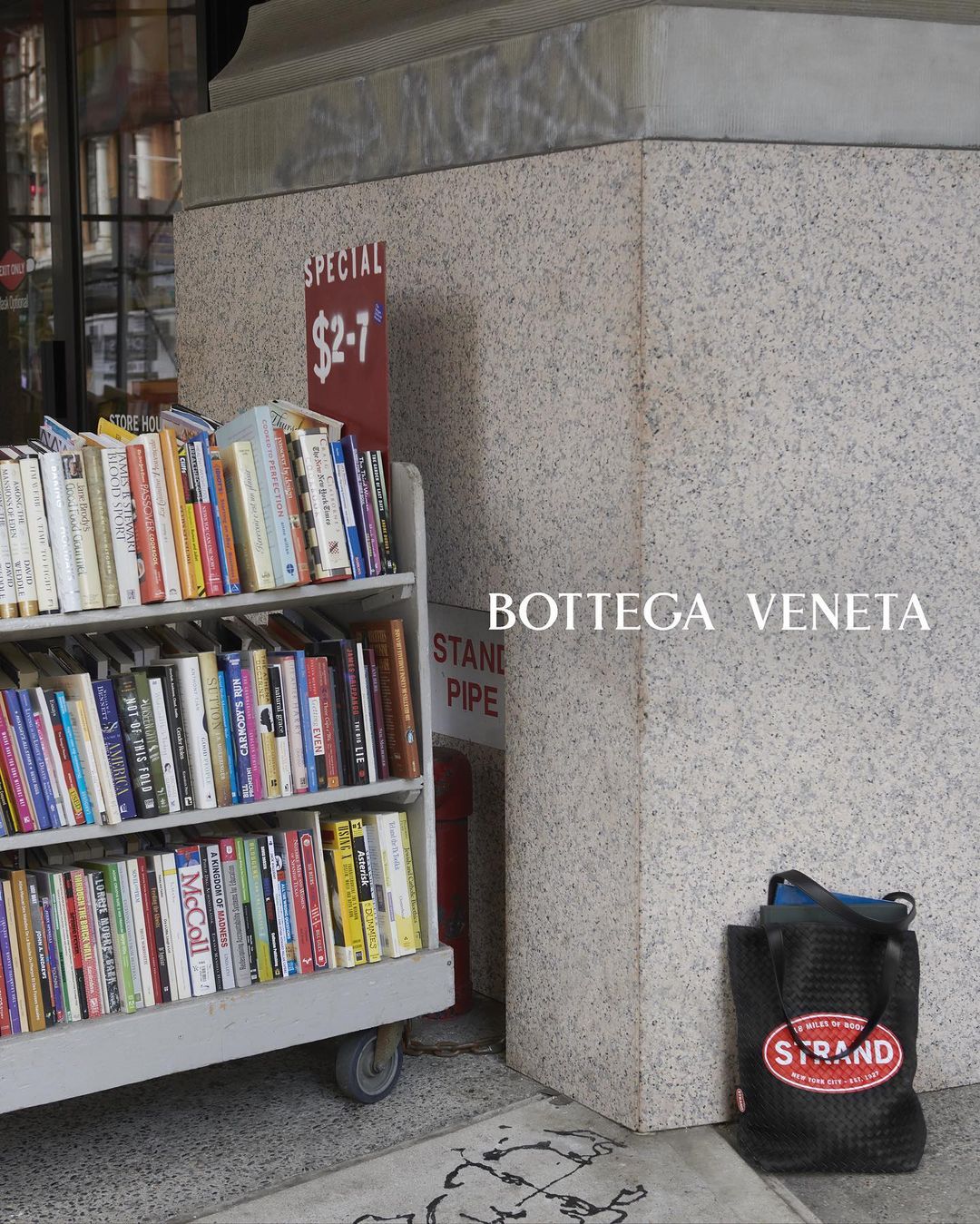 Outdoor book display rack with various books near a gray concrete column and a black tote bag with the Strand Bookstore logo.
