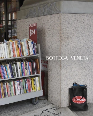 Outdoor book display rack with various books near a gray concrete column and a black tote bag with the Strand Bookstore logo.