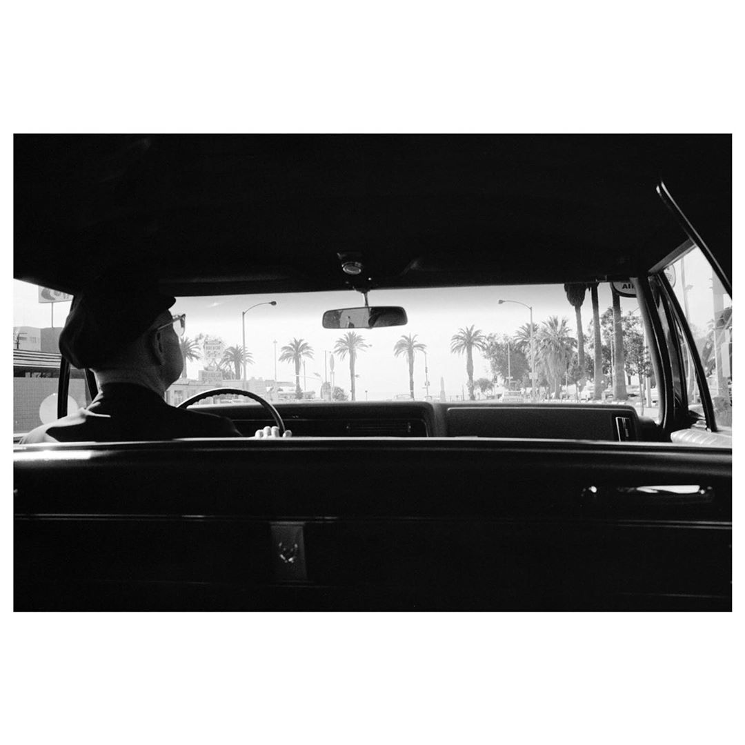 View from inside classic car with an elderly man driving, palm trees and buildings in the background in black and white.
