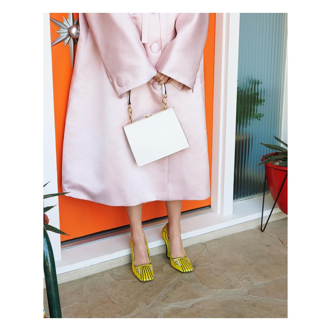 Fashion shoot features a woman in a pink coat and yellow shoes holding a white purse, posing in front of an orange and white door.