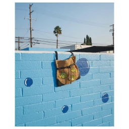 Woven straw handbag with embroidered fish patches hanging on a bright blue painted brick wall under a clear sky.