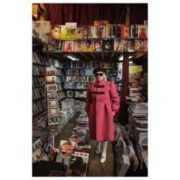 A person wearing a pink coat and black beret stands in a bookstore filled with shelves and stacks of magazines.
