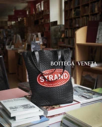 Black woven leather tote bag with Strand Bookstore logo resting on stacks of books inside a bookstore.