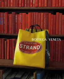 A yellow and black Strand Bookstore tote bag positioned in front of a floor-to-ceiling bookshelf filled with red books.