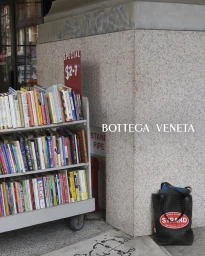 A rolling book cart filled with colorful paperbacks next to a granite building pillar with a black tote bag on the sidewalk.
