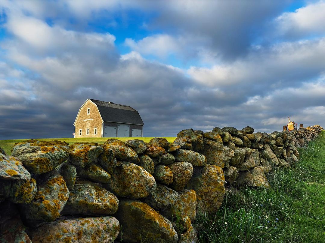Rural landscape with a historic stone wall, a classic barn, and a tractor in the background under a bright, partly cloudy sky.